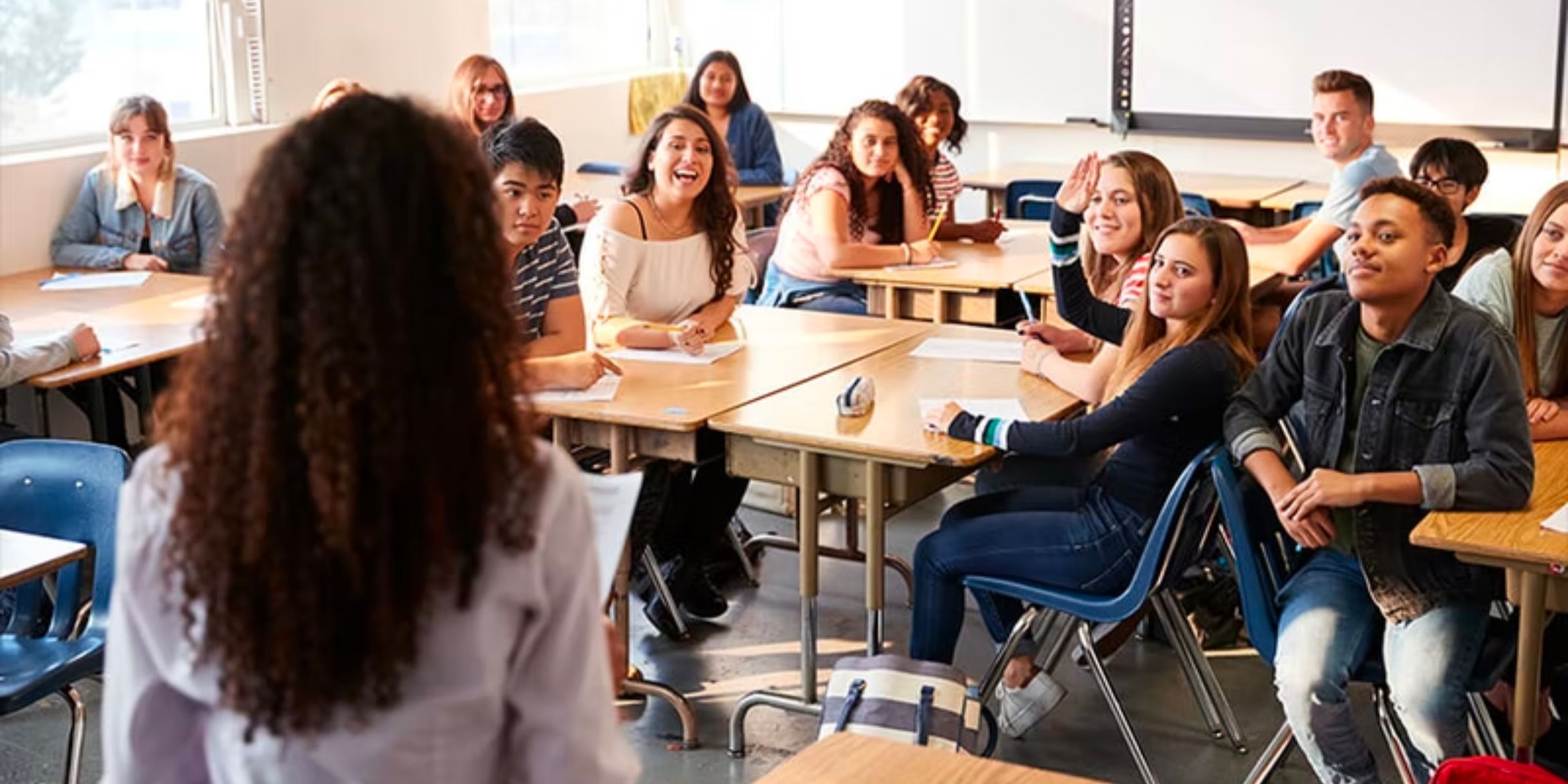 high school students at their desks smiling as they listen to a teacher 