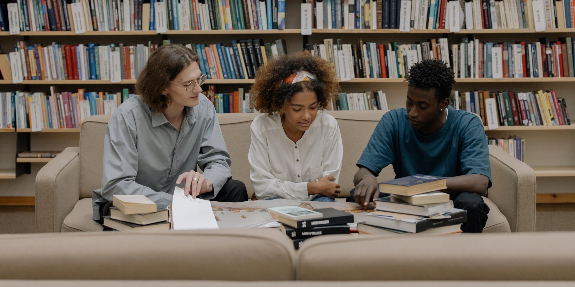 three students sitting on a couch in a library and talking as they look at books on a table together