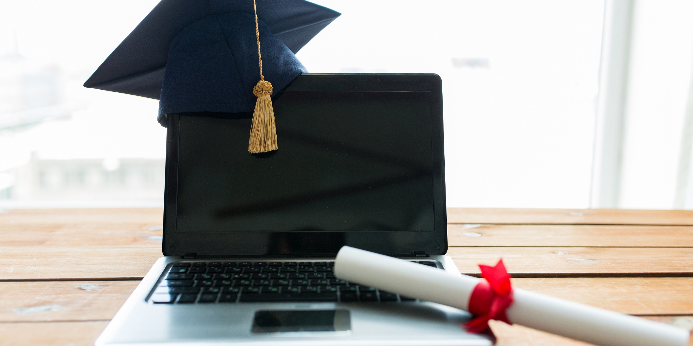 an open laptop with a graduation cap resting on a top corner and a rolled up diploma on the keyboard
